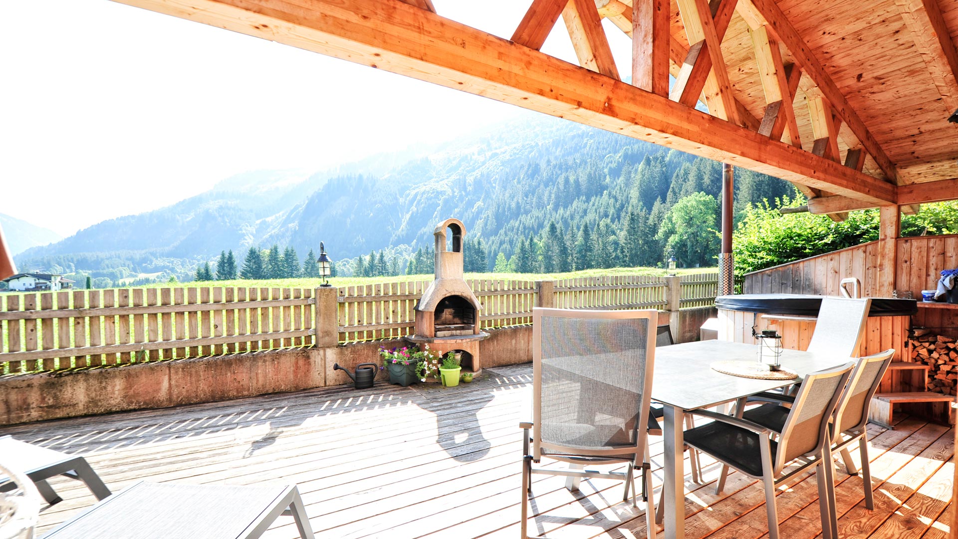 Überdachte Holzterrasse mit Grill, Gartenmöbeln und Bergblick im Sommer.